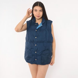 May include: A sleeveless navy blue puffer vest featuring a high collar, a front button closure with silver-toned metal snaps, and two front patch pockets. The interior lining of the collar is a contrasting light blue fabric.
