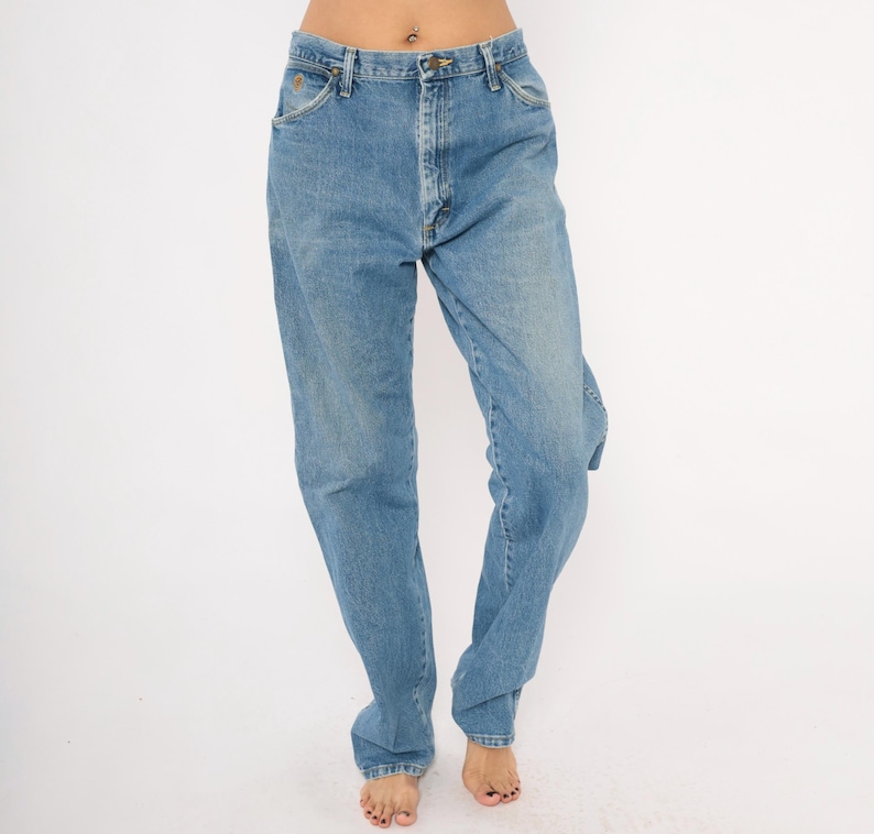 Pu&ograve; includere: Jeans di denim blu chiaro con un classico design a cinque tasche. I jeans hanno una vestibilit&agrave; a gamba dritta e una chiusura con bottoni e cerniera. Il denim ha un lavaggio sbiadito, che conferisce loro un aspetto vintage. I jeans sono indossati da una persona.