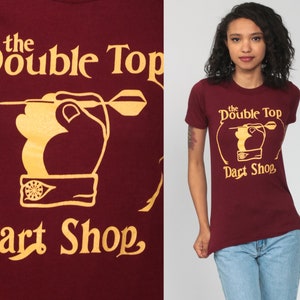 May include: A maroon t-shirt with a gold graphic of a hand throwing a dart at a dartboard. The graphic says "the Double Top Dart Shop".