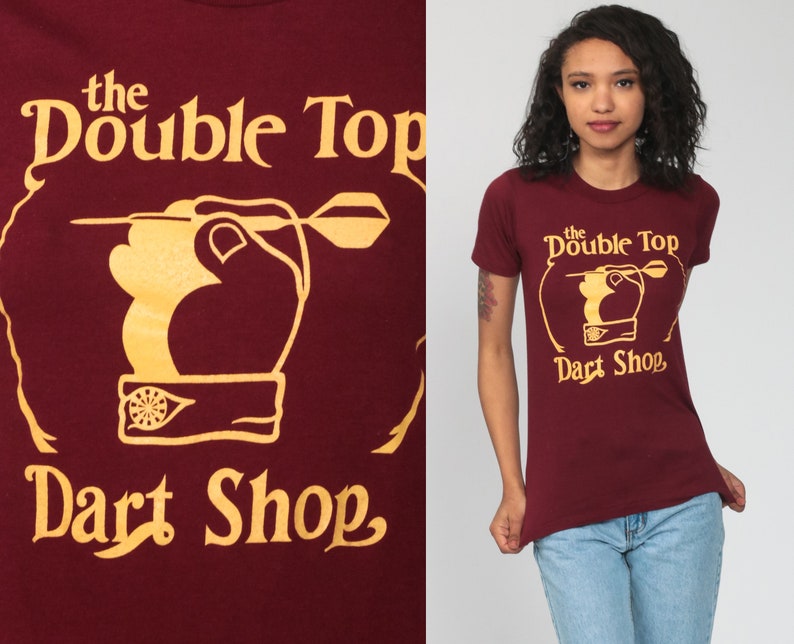 May include: A maroon t-shirt with a gold graphic of a hand throwing a dart at a dartboard. The graphic says "the Double Top Dart Shop".