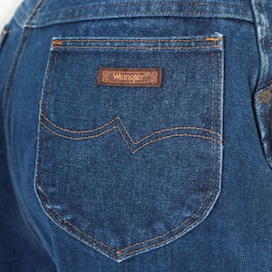 May include: Close-up of a dark blue denim jean pocket with orange stitching. A brown leather patch with the "Wrangler" logo is centered above the pocket. The pocket has a distinctive "W" shaped design.