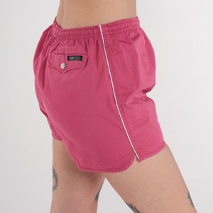 May include: Bright pink athletic shorts with white piping along the seams. These shorts feature an elasticated waistband and a small pocket on the back with a button. Made from a lightweight material.