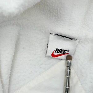 Puede incluir: Primer plano de una chaqueta de forro polar blanca con el parche del logo de Nike. El logo presenta el nombre de la marca en negro y un swoosh rojo. La chaqueta tiene un cord&oacute;n y un aspecto suave y texturizado.