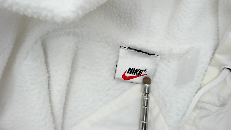 Puede incluir: Primer plano de una chaqueta de forro polar blanca con el parche del logo de Nike. El logo presenta el nombre de la marca en negro y un swoosh rojo. La chaqueta tiene un cord&oacute;n y un aspecto suave y texturizado.