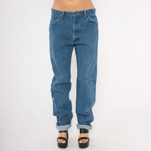 May include: Blue denim jeans with a classic fit. The jeans have a button and zipper closure, front and back pockets, and are cuffed at the ankles. The model is wearing black platform sandals. The jeans are a medium wash.