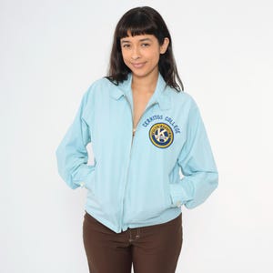 Puede incluir: Una chaqueta azul claro con cierre de cremallera y un parche en el pecho izquierdo que dice "Cerritos College" con un logotipo circular.