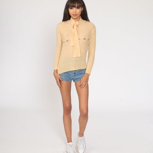 May include: A pale yellow knitted sweater with a tie at the neck and floral embroidery. The sweater has long sleeves and a ribbed texture.