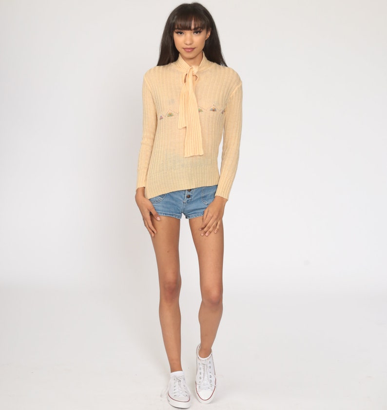 May include: A pale yellow knitted sweater with a tie at the neck and floral embroidery. The sweater has long sleeves and a ribbed texture.