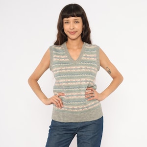 May include: A gray V-neck sweater vest with a patterned design in light blue, pink, and white. The sleeveless vest is worn by a person wearing blue jeans.