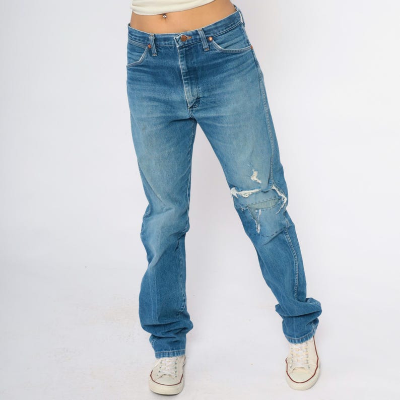 May include: Faded blue denim jeans with a distressed knee detail. The jeans have a classic five-pocket design and a straight leg fit. The model is wearing white canvas trainers.