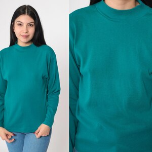 Pode incluir: Camisa de manga comprida verde-azulado com gola alta. A camisa &eacute; feita de um material macio e &eacute; de cor lisa. A modelo est&aacute; usando jeans azuis. A camisa &eacute; de cor verde-azulado lisa.