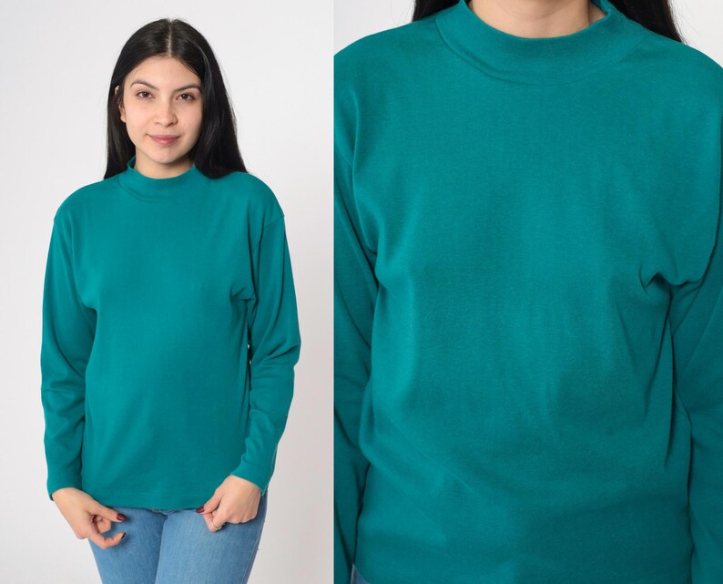 Pode incluir: Camisa de manga comprida verde-azulado com gola alta. A camisa &eacute; feita de um material macio e &eacute; de cor lisa. A modelo est&aacute; usando jeans azuis. A camisa &eacute; de cor verde-azulado lisa.