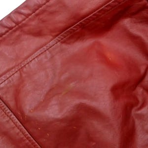 May include: Close-up of a section of a red leather jacket. The image shows the shoulder and upper chest area, with visible stitching and seams. The leather has a slightly glossy finish and some minor imperfections.