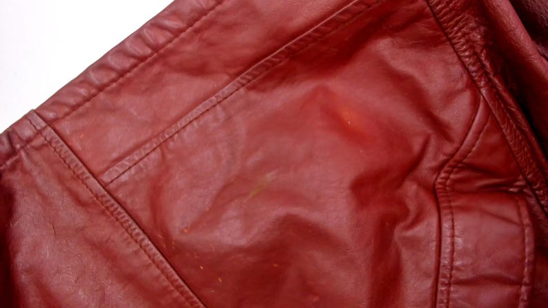 May include: Close-up of a section of a red leather jacket. The image shows the shoulder and upper chest area, with visible stitching and seams. The leather has a slightly glossy finish and some minor imperfections.