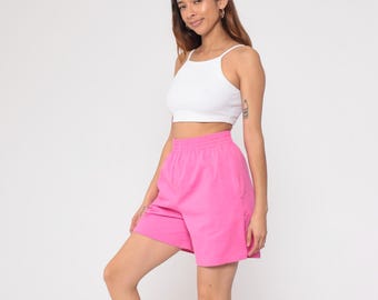 Puede incluir: Un top corto blanco y unos shorts rosa brillante. Los shorts tienen cintura con cordón y bolsillos. El conjunto es casual y deportivo, perfecto para el clima cálido. La modelo sonríe y mira a la cámara.