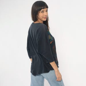 May include: Dark navy blue three-quarter sleeve shirt with a colorful graphic design on the upper right side. The shirt is paired with light blue jeans. The shirt has a relaxed fit and a v-neck.
