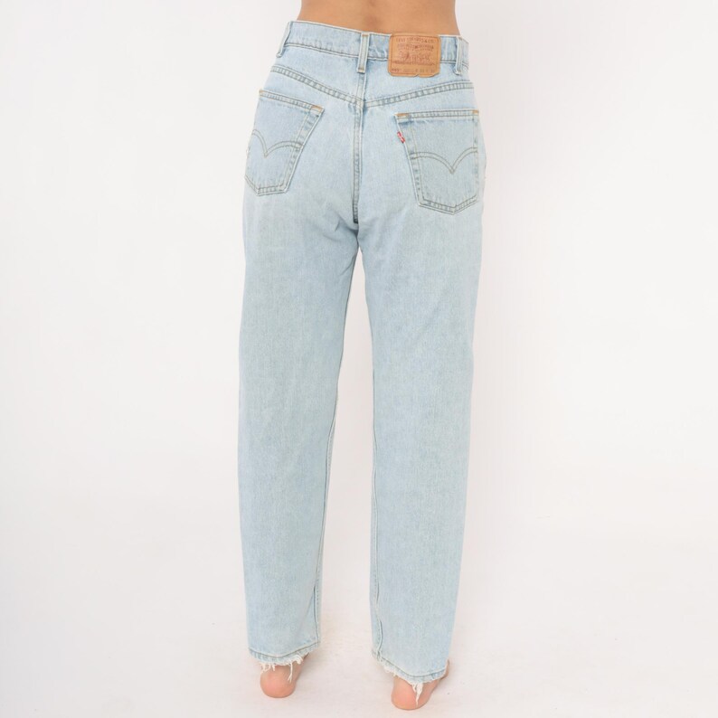 May include: Light blue denim jeans with a high waist and straight leg. The jeans have classic five-pocket styling, a leather patch, and a red tab on the back pocket. The hem is frayed. The jeans are shown from the back.