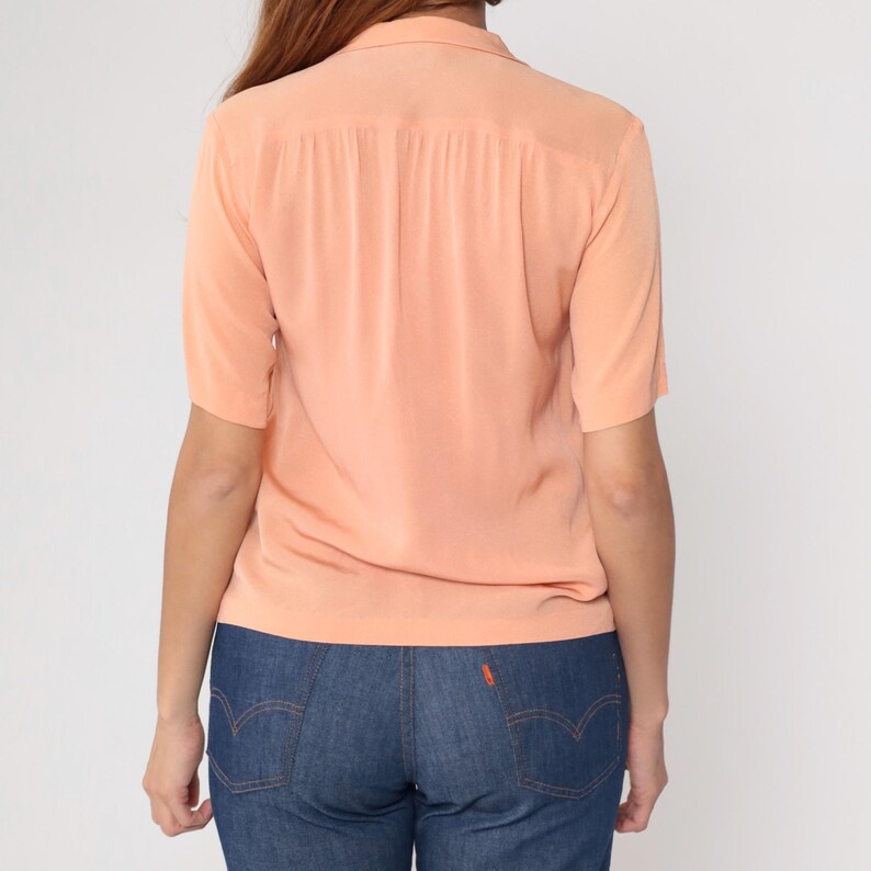 Puede incluir: Blusa melocot&oacute;n de manga corta con cuello. La parte trasera de la blusa presenta pliegues verticales. La blusa se combina con vaqueros azules.