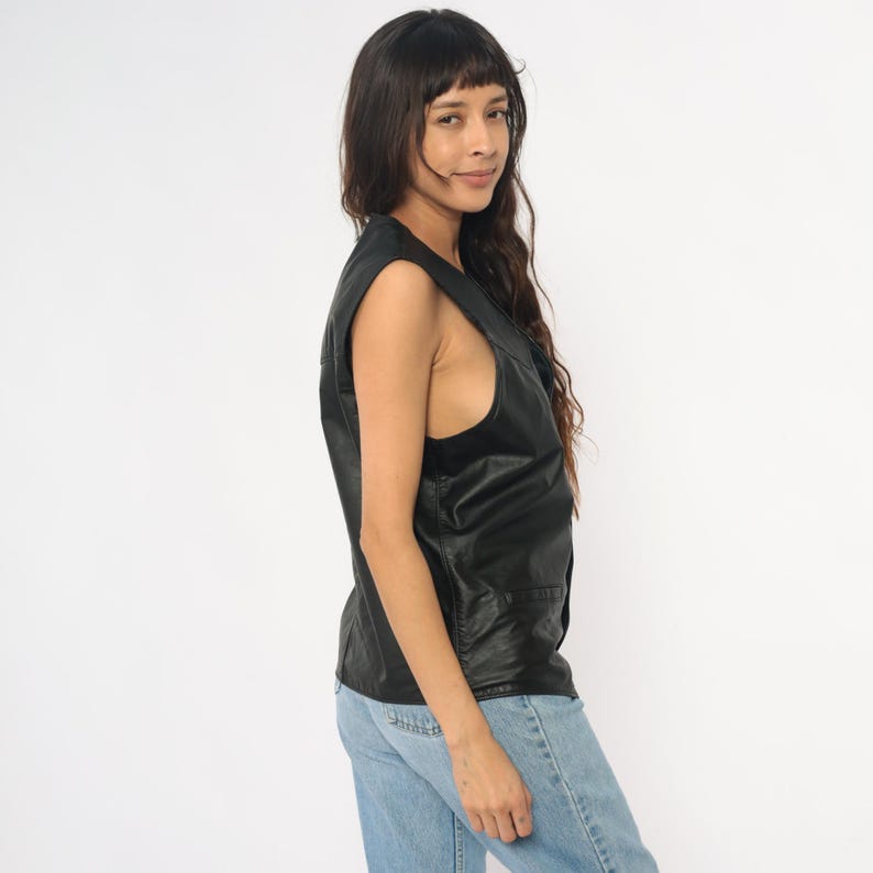 May include: A black leather vest with a V-neck and two front pockets. The vest is sleeveless and appears to be a vintage style. The model is wearing blue jeans.