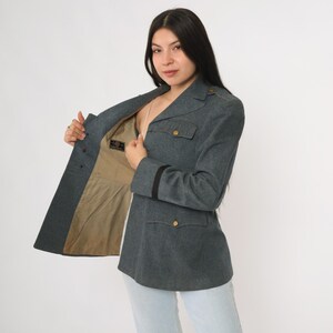 K&ouml;nnte beinhalten: Graue Jacke im Milit&auml;rstil mit goldenen Kn&ouml;pfen und schwarzen Streifen an den &Auml;rmeln. Die Jacke hat zwei Vordertaschen und eine taillierte Passform. Das Innenfutter ist beige.
