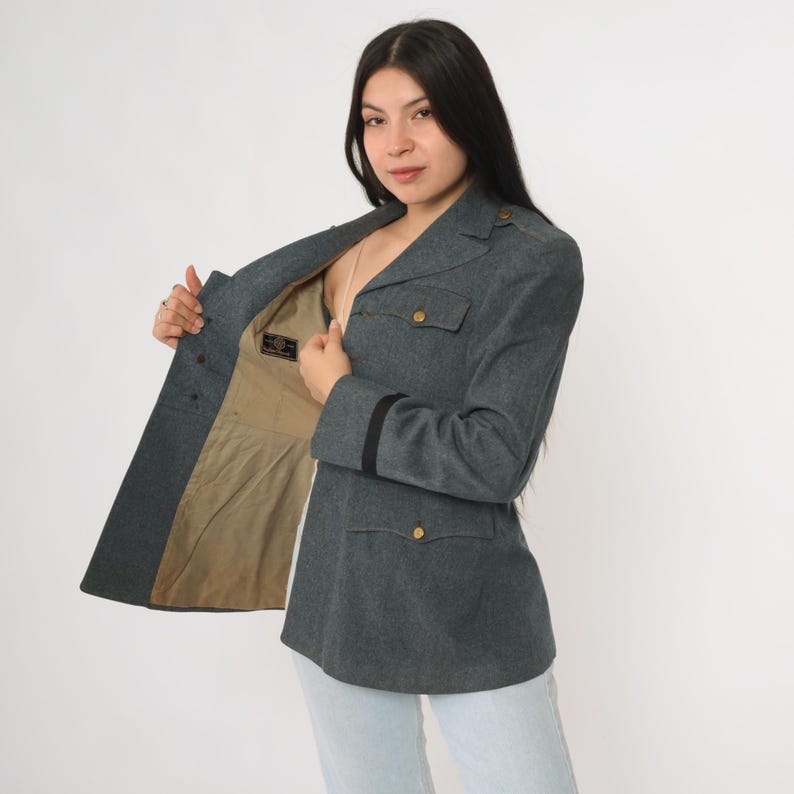 K&ouml;nnte beinhalten: Graue Jacke im Milit&auml;rstil mit goldenen Kn&ouml;pfen und schwarzen Streifen an den &Auml;rmeln. Die Jacke hat zwei Vordertaschen und eine taillierte Passform. Das Innenfutter ist beige.