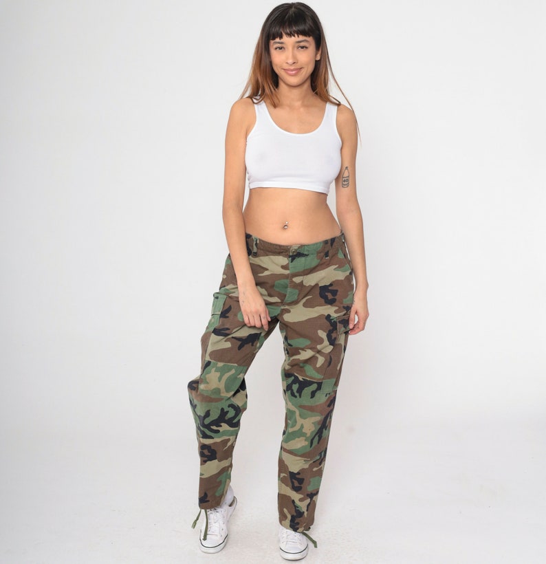 May include: A white tank top and camouflage cargo pants. The pants feature a green, brown, and black pattern. The outfit is paired with white sneakers. The overall style is casual and utilitarian.