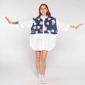 May include: A white button-down shirt with a blue and red patchwork design featuring hearts and squares. The shirt has short sleeves and a relaxed fit.