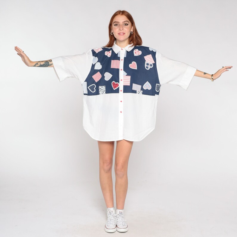 May include: A white button-down shirt with a blue and red patchwork design featuring hearts and squares. The shirt has short sleeves and a relaxed fit.