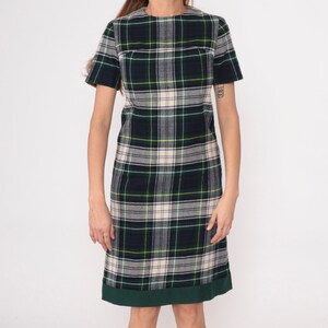 May include: A short-sleeved plaid dress in shades of green, navy, yellow, and cream. The dress features a classic plaid pattern and a solid green hemline. The dress is knee-length and has a simple, straight silhouette.