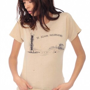 May include: Tan short-sleeved t-shirt with a graphic print. The print features a black and white illustration of a structure and the text "ST. JOHN NEUMANN". The shirt is made of a soft, lightweight material.
