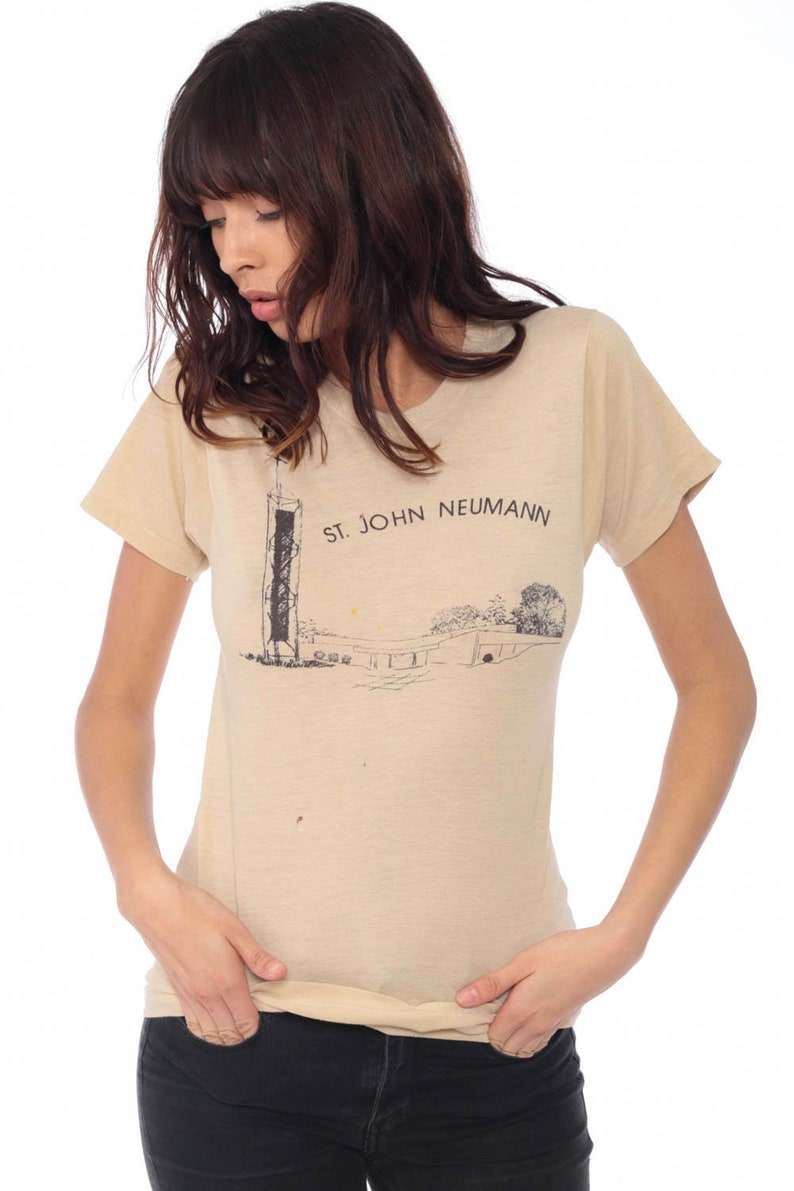 May include: Tan short-sleeved t-shirt with a graphic print. The print features a black and white illustration of a structure and the text "ST. JOHN NEUMANN". The shirt is made of a soft, lightweight material.