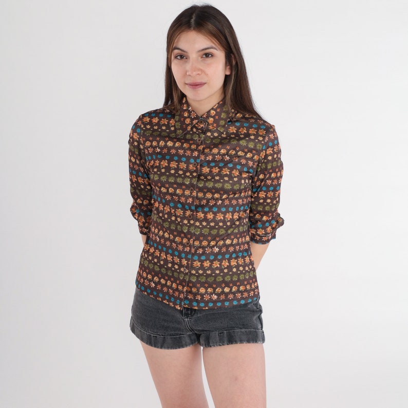 May include: A brown, long-sleeved shirt with a colorful floral pattern. The shirt has a button-up collar and a fitted silhouette.