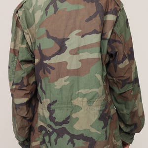 May include: A long-sleeved field jacket featuring a woodland camouflage pattern in shades of olive green, brown, and black. The jacket includes a fold-down collar, shoulder epaulets, and a cinched waist detail, providing a structured fit.
