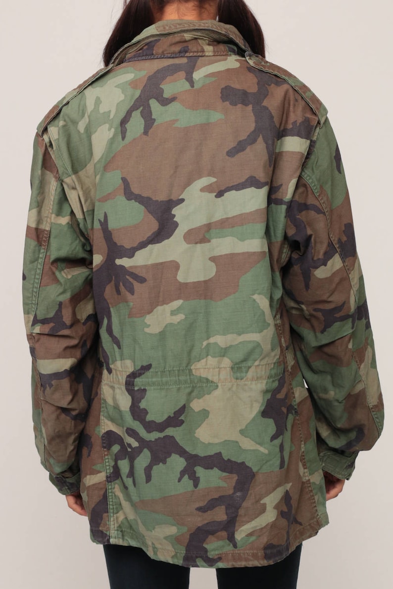 May include: A long-sleeved field jacket featuring a woodland camouflage pattern in shades of olive green, brown, and black. The jacket includes a fold-down collar, shoulder epaulets, and a cinched waist detail, providing a structured fit.