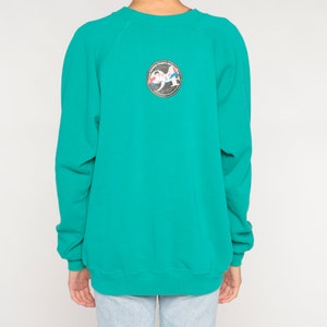 May include: Teal-colored sweatshirt with a crew neck and long sleeves. A circular patch on the back features a design with the number "94" and other graphic elements. The sweatshirt is made of a soft material.