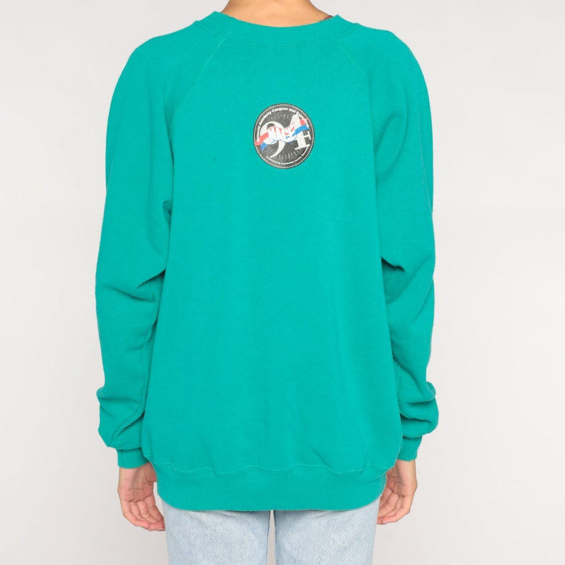 May include: Teal-colored sweatshirt with a crew neck and long sleeves. A circular patch on the back features a design with the number "94" and other graphic elements. The sweatshirt is made of a soft material.
