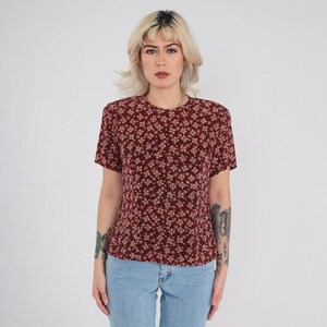 May include: A burgundy short-sleeve blouse with a repeating floral pattern in tan and cream. The blouse has a round neckline and is paired with light blue denim jeans. The model is standing in front of a white background.