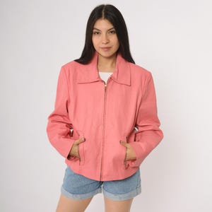 Könnte beinhalten: Korallfarbene Jacke mit Kragen und durchgehendem Reißverschluss. Die Jacke hat zwei Vordertaschen mit Reißverschlüssen und lange Ärmel. Das Model trägt Jeansshorts. Die Jacke ist aus einem glatten, einfarbigen Stoff gefertigt.