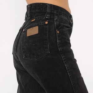 May include: Black Wrangler jeans with a high waist and a classic fit. The jeans feature a brown leather patch and copper rivets. The Wrangler logo is visible on the back pocket and waistband. A timeless denim staple.