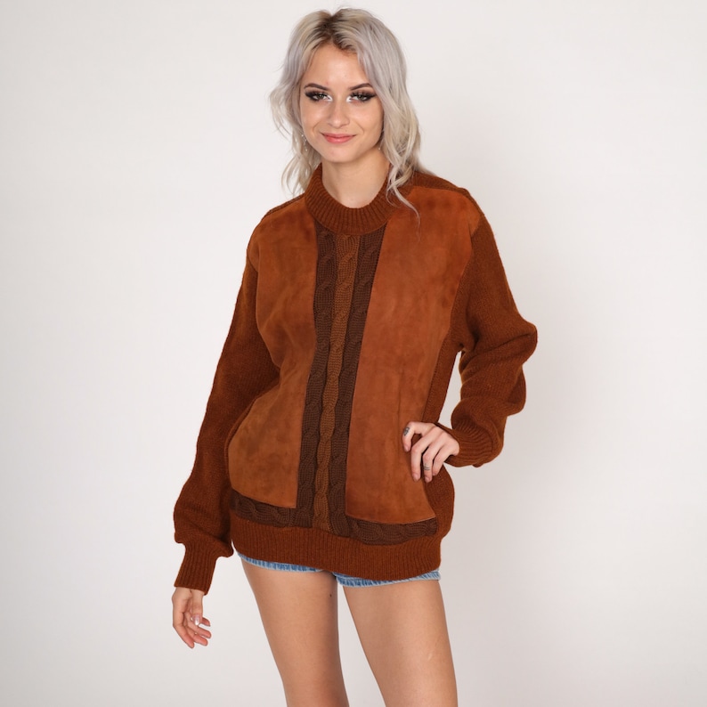 May include: A brown sweater with a suede panel on the front. The sweater has a cable knit design and a crew neck.