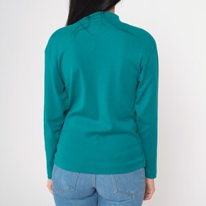 Pode incluir: Camisa de manga comprida verde-azulado com gola alta. A camisa &eacute; combinada com jeans azul claro. A vista traseira da camisa mostra o decote e o ajuste da pe&ccedil;a. O tecido parece ser um tecido de malha macia.