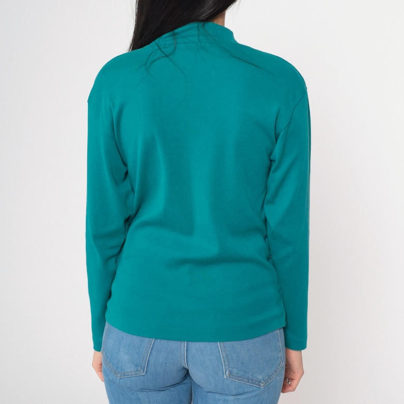 Pode incluir: Camisa de manga comprida verde-azulado com gola alta. A camisa &eacute; combinada com jeans azul claro. A vista traseira da camisa mostra o decote e o ajuste da pe&ccedil;a. O tecido parece ser um tecido de malha macia.