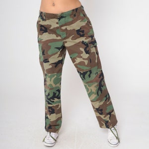 May include: Camouflage cargo pants in shades of green, brown, and black. The pants feature multiple pockets and a straight leg design. The pants are worn with white sneakers.