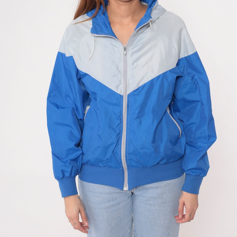 May include: A blue and silver windbreaker jacket with a zipper closure and a hood. The jacket has a color block design with a silver panel on the front and blue panels on the sleeves and back.