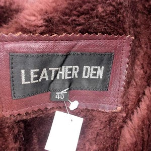 May include: Close-up of a burgundy leather label with the text "LEATHER DEN" in white. The label is stitched onto a burgundy leather garment with a faux fur lining. A tag with the number "40" is attached.