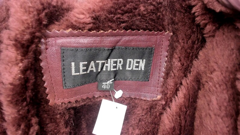 May include: Close-up of a burgundy leather label with the text "LEATHER DEN" in white. The label is stitched onto a burgundy leather garment with a faux fur lining. A tag with the number "40" is attached.