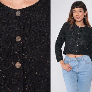 Puede incluir: Top negro de manga larga con estampado floral y botones redondos. El top es corto y tiene cuello redondo. Los botones son de color met&aacute;lico. La modelo lleva vaqueros azul claro.