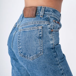 May include: Light blue denim jeans with a classic five-pocket design. The jeans feature a brown leather patch on the back and a small logo detail on the front pocket. The jeans are a medium wash and have a straight leg fit.