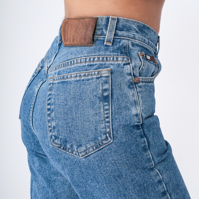 May include: Light blue denim jeans with a classic five-pocket design. The jeans feature a brown leather patch on the back and a small logo detail on the front pocket. The jeans are a medium wash and have a straight leg fit.