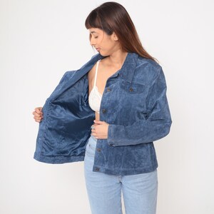 May include: A blue suede jacket with a button-up front and two chest pockets. The jacket is open, revealing a blue satin lining. The person is wearing light blue jeans and a white bra.
